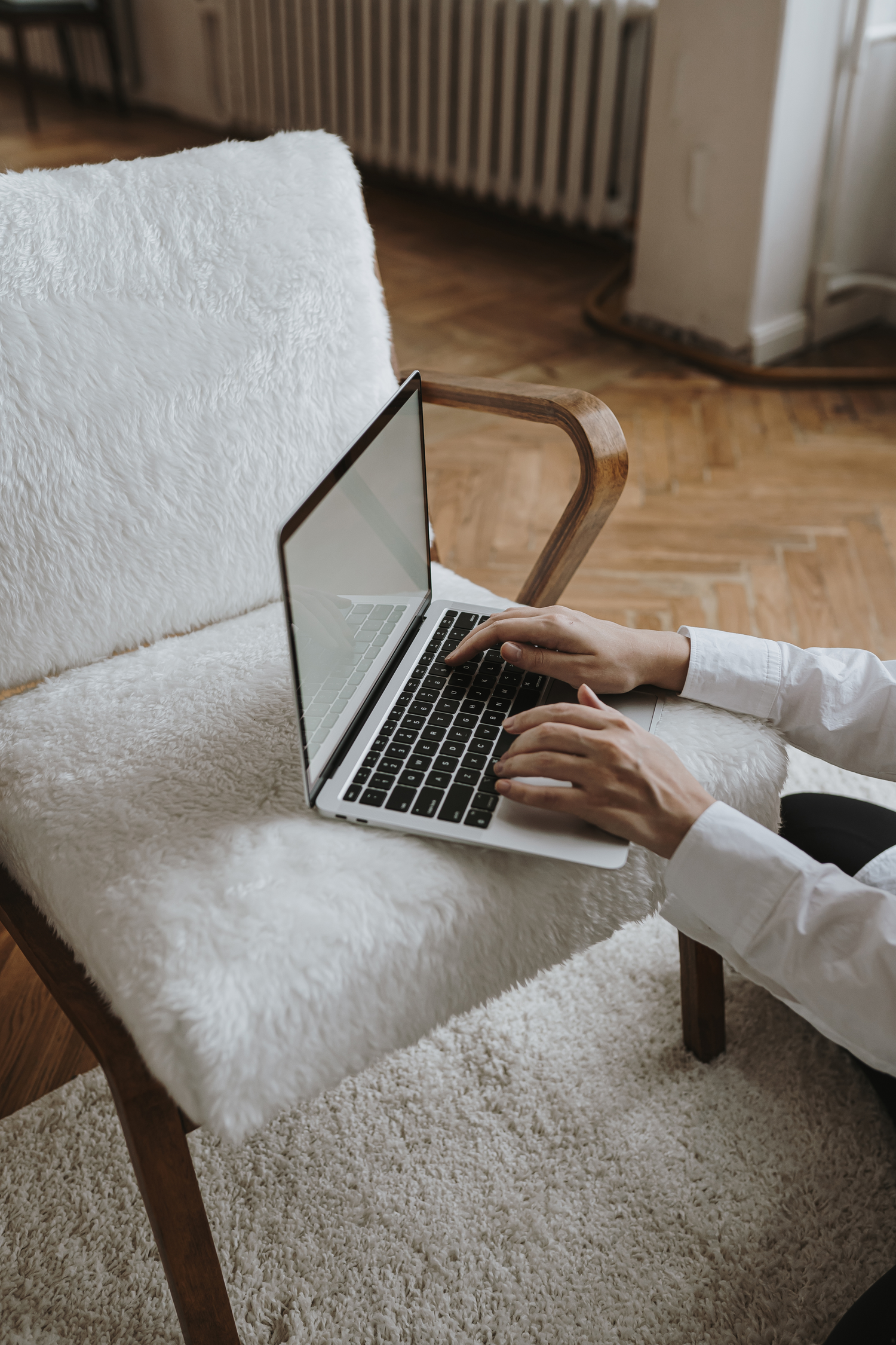 Model sitting on floor typing on a laptop that's sitting on a white fuzzy chair in a neutral apartment space