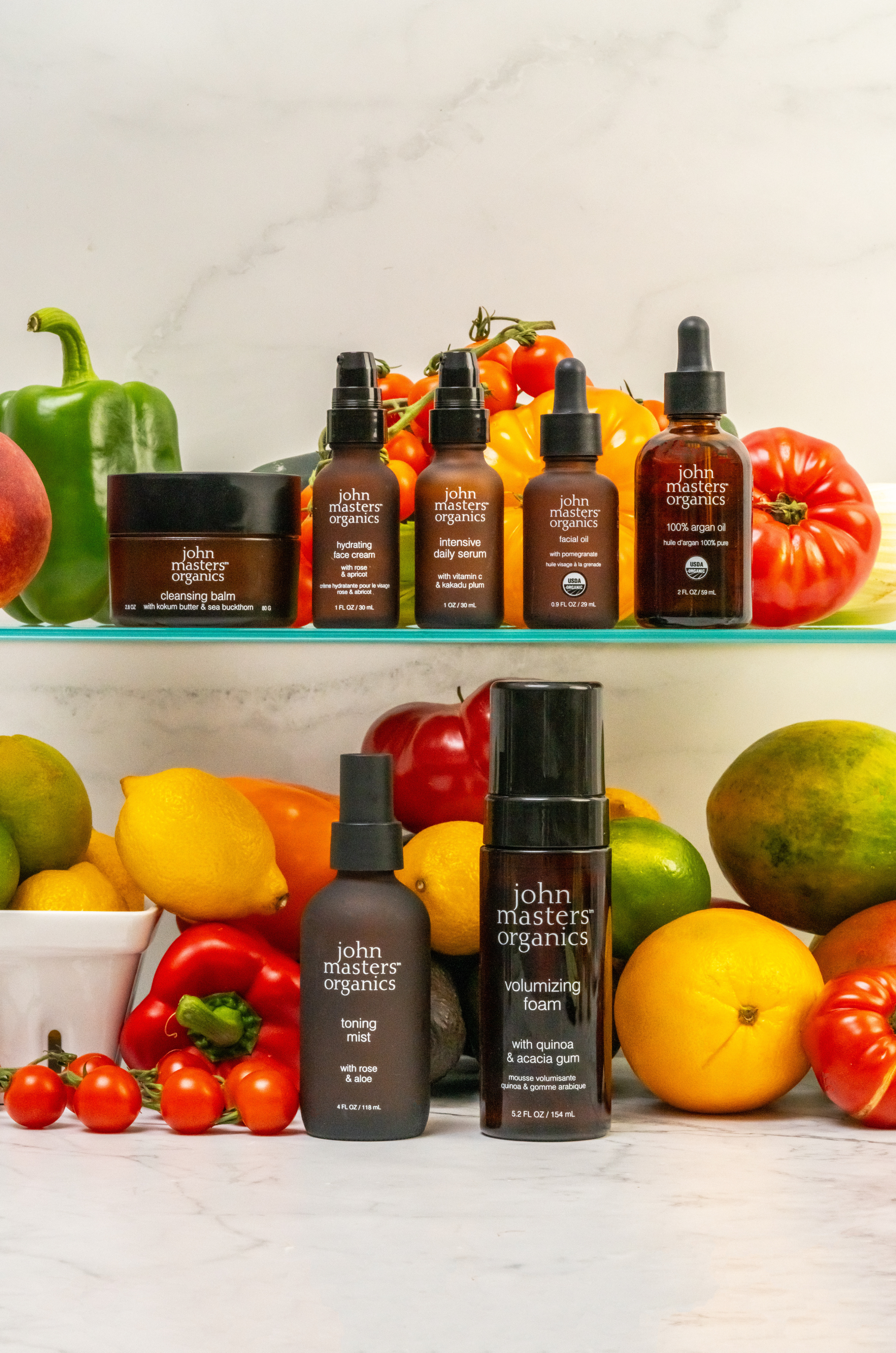 John Masters Organics Cleansing Balm, Hydrating Face Cream, Intensive Daily Serum, Facial Oil, 100% Argan Oil, Toning Mist, and Volumizing Foam on shelves surrounded by vegetables