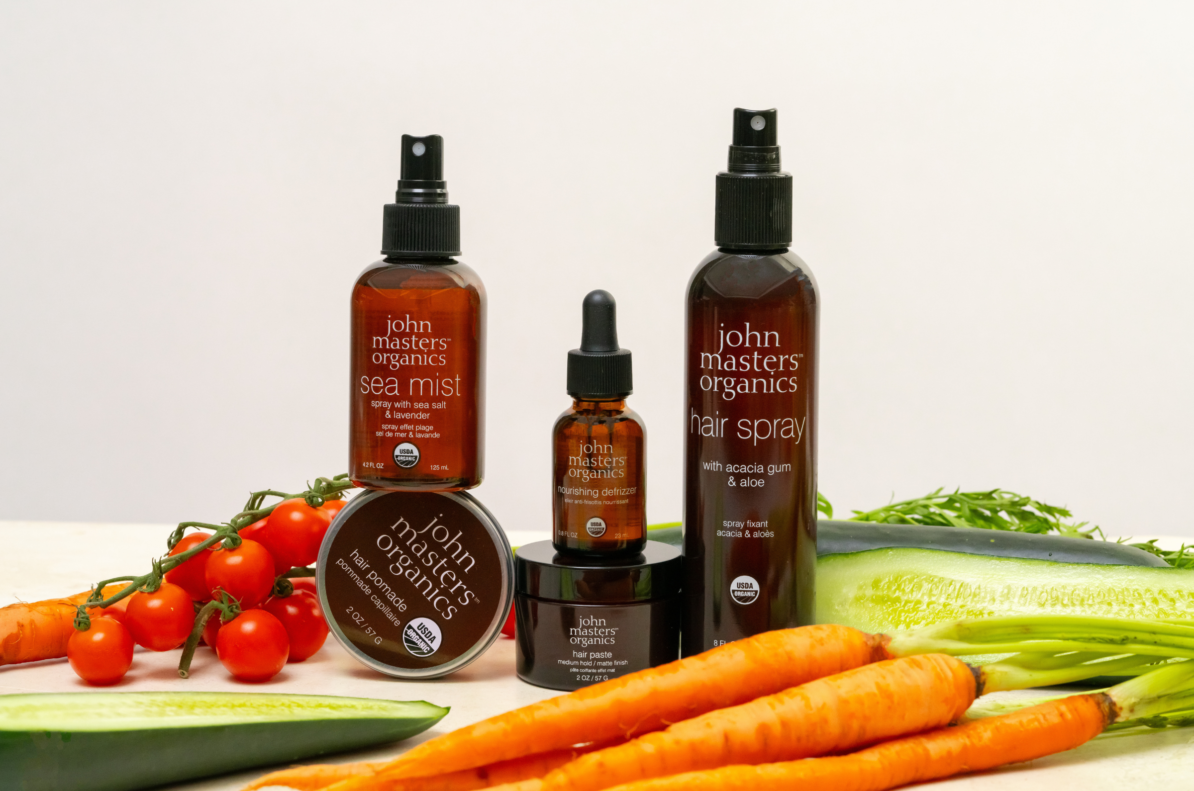 John Masters Organics Sea Mist, Hair Pomade, Nourishing Defrizzer, Hair Paste, and Hair Spray surrounded by tomatoes, cucumbers and carrots 