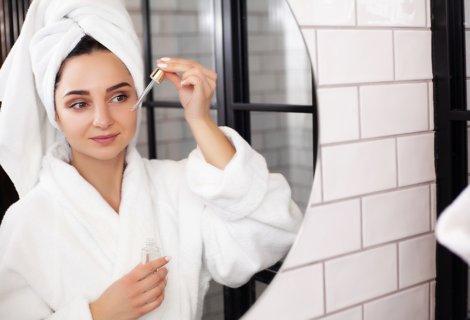 Mirror reflection of a model applying serum to face while standing in the bathroom