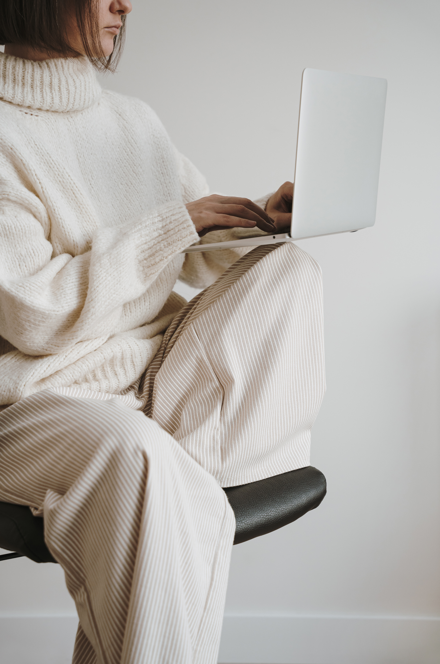 Model sitting on a swivel chair in an all neutral outfit typing on a laptop
