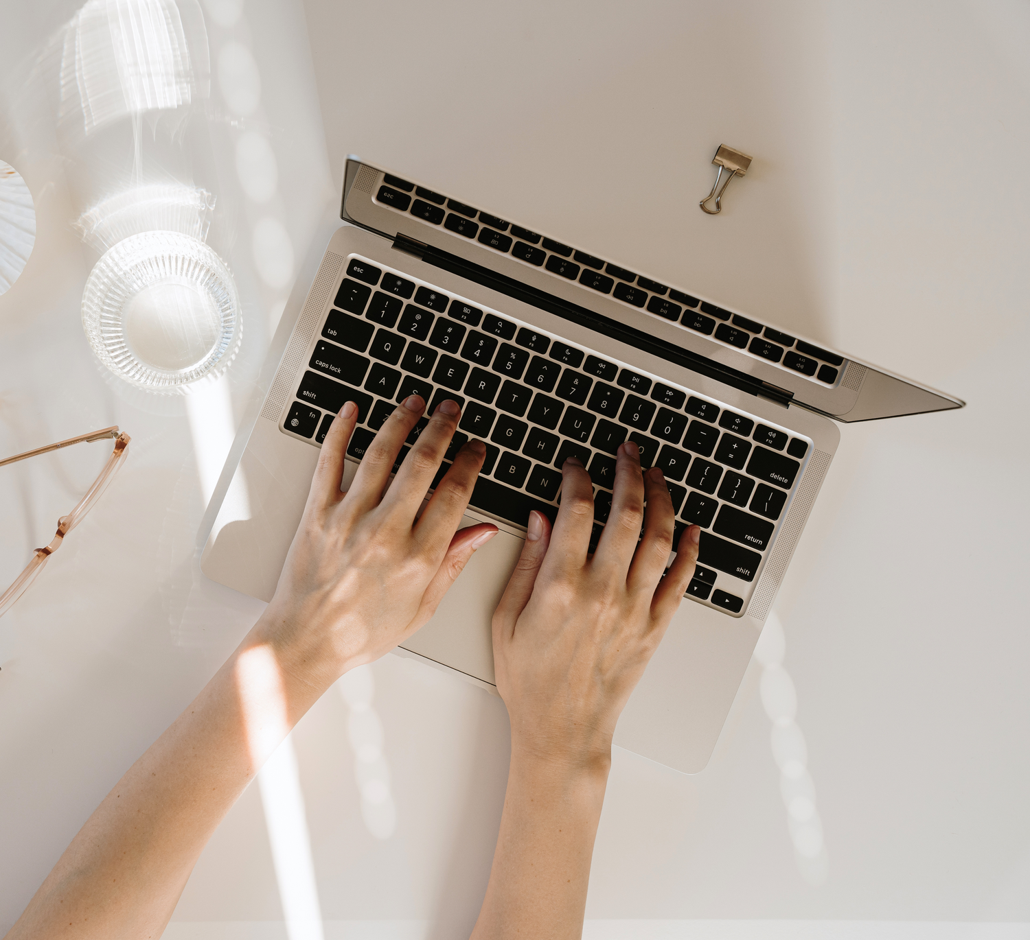 Model hands typing on a laptop that is surrounded by reading glasses, a glass of water and a paper clip on a white table