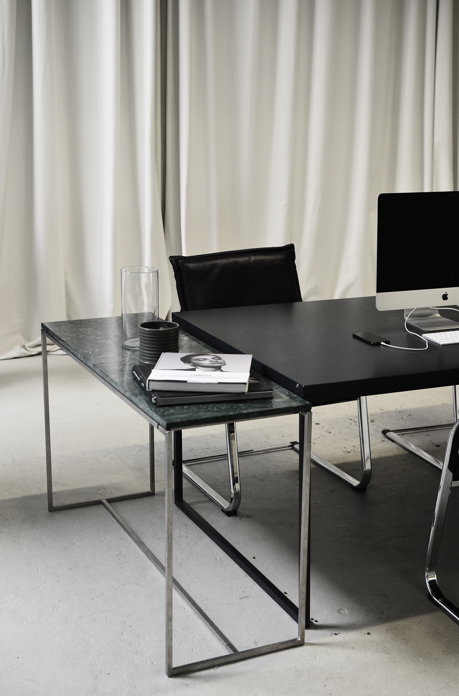 Black desk with Apple monitor, iPhone plugged in charging, coffee table books, glass case and pottery jar on it